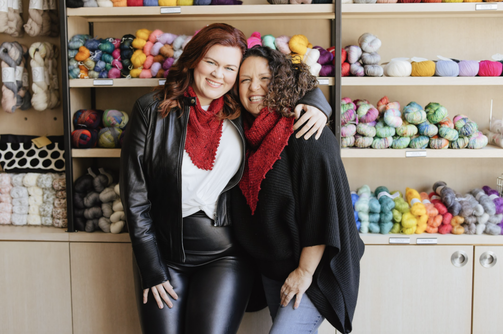 Crochet and Knit designer Michele Costa of 144 Stitches and Indie Yarn Dyer Stephanie of Asylum fibers hugging and wearing knitted shawls.