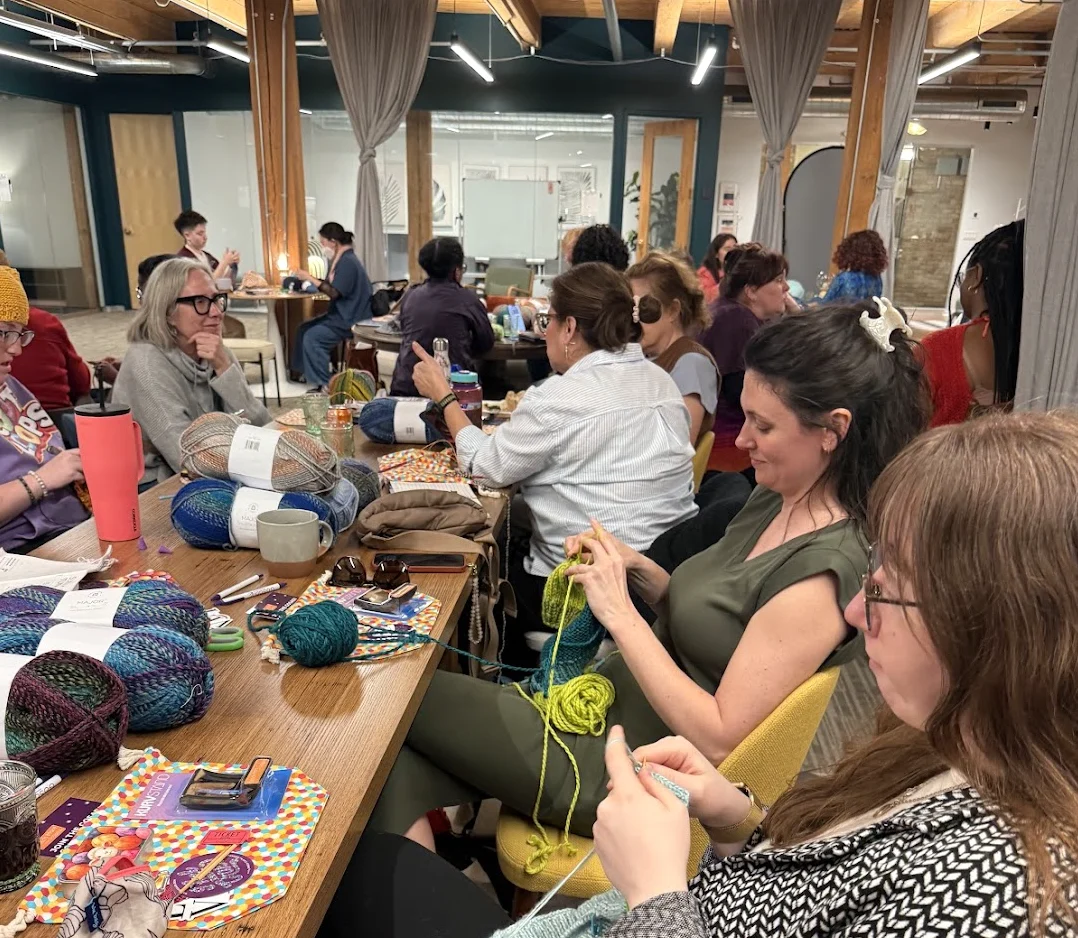 Makers at a stitching circle meetup with knitting needles and yarn projects, gathered around a table at a GoSadi knitting community event