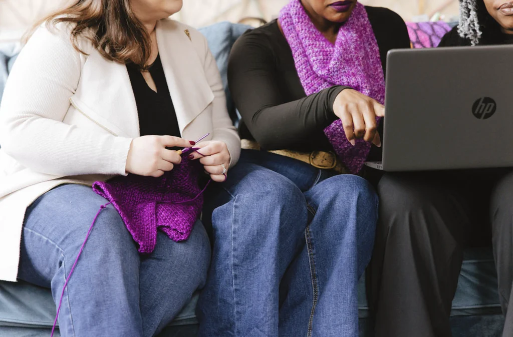 Crochet designers working on laptop with colorful yarn and projects, including GoSadi purple shawls.