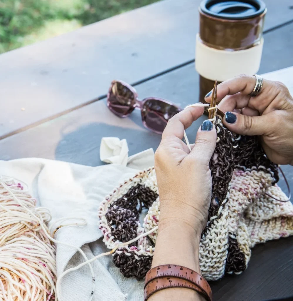 Designer knitting with yarn, measuring tape, knitting needles, and notions needed to calculate yarn amounts for custom patterns.