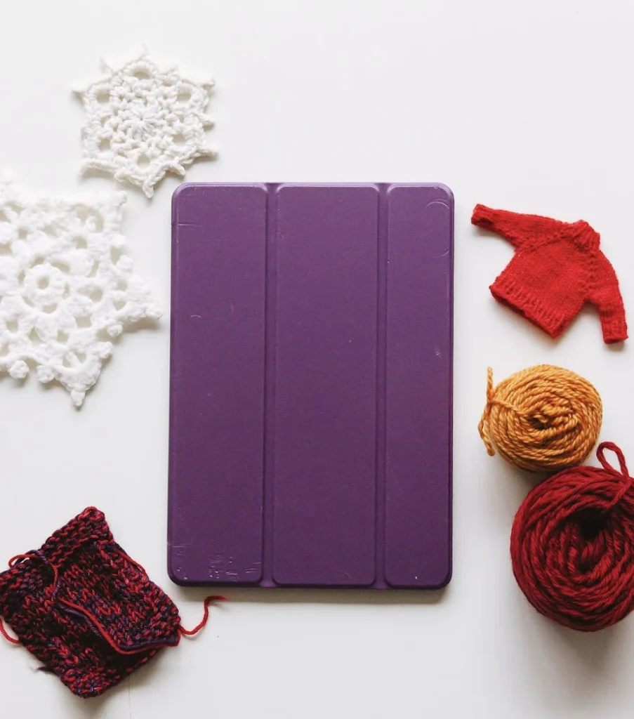 Designer's flat lay showing holiday knitting projects next to a purple tablet, featuring white crochet snowflakes, a red knit sweater, and colorful yarn for seasonal pattern planning.