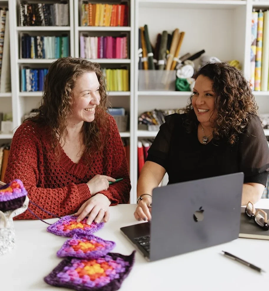 Tamara Kelly of Moogly Blog and Michele Costa of GoSadi update her Etsy account on a laptop in front of a yarn wall.